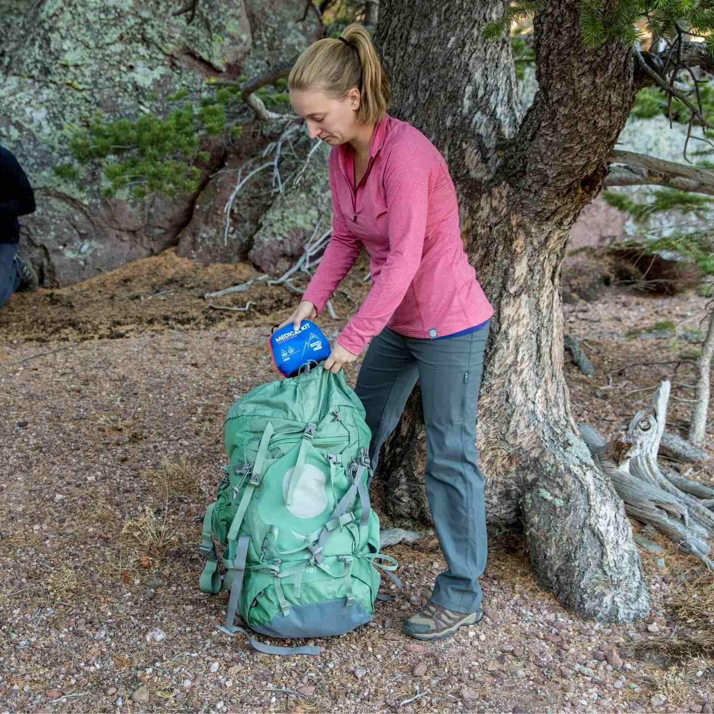 Mountain Series Medical Kit - Hiker woman in pink shirt pulling kit from green back in front of tree