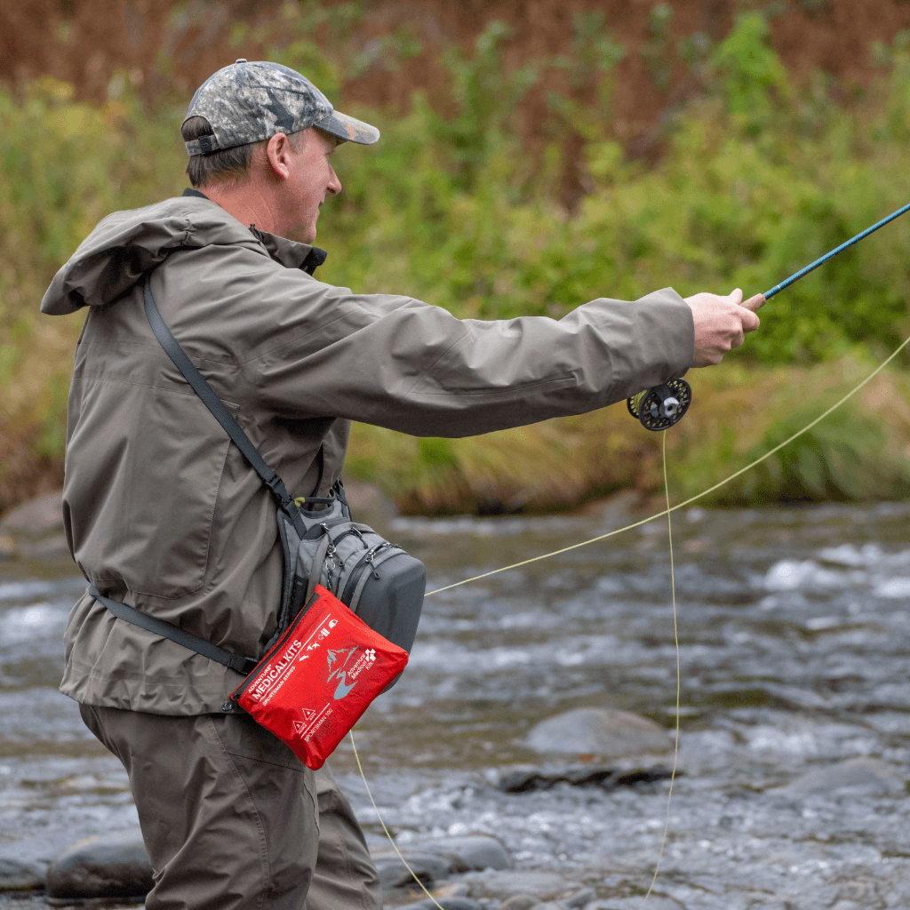 A man fly fishing in a river, wearing a gray hooded jacket, gray pants, and a camouflage cap, with a red and gray fly fishing bag hanging from his shoulder.