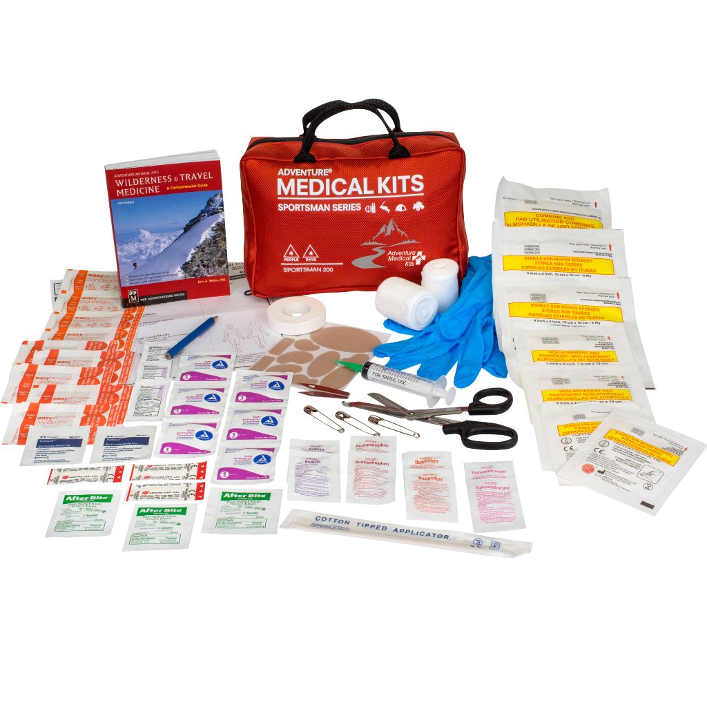 A comprehensive first aid and wilderness medical kit set displayed on a clean white background, featuring a red zippered 'Adventure Medical Kits Sportman Series' bag, a Wilderness Travel Medicine book, blue gloves, gauze, bandages, scissors, tweezers, and multiple individually wrapped medical supplies.