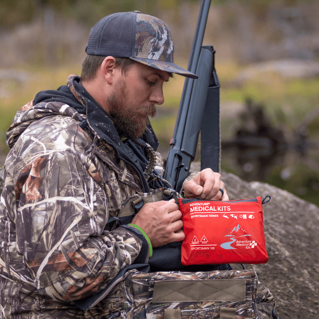 A man in camouflage hunting clothing holds a red first aid medical kit pouch while standing outdoors near a rifle.