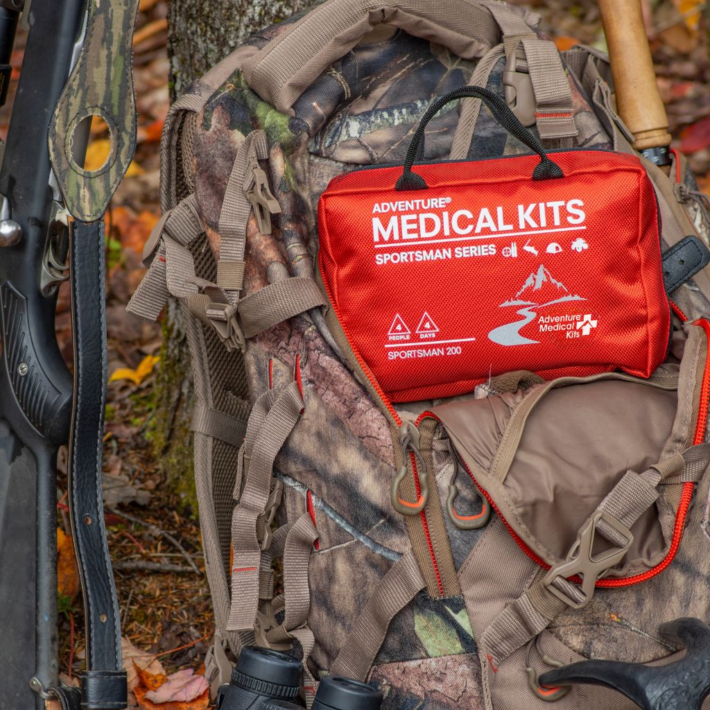 A red Adventure Medical Kits Sportsman Series first aid kit pouch is placed on top of a camouflage backpack outdoors, with a rifle and other outdoor gear visible around it.
