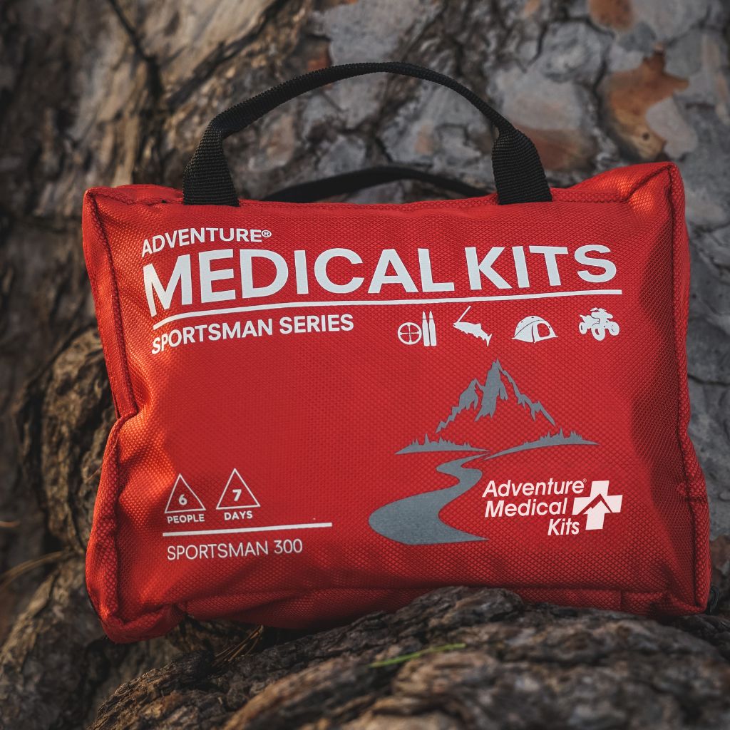 A red Adventure Medical Kits Sportsman Series 300 first aid kit bag with black handles, placed on a textured tree trunk background.