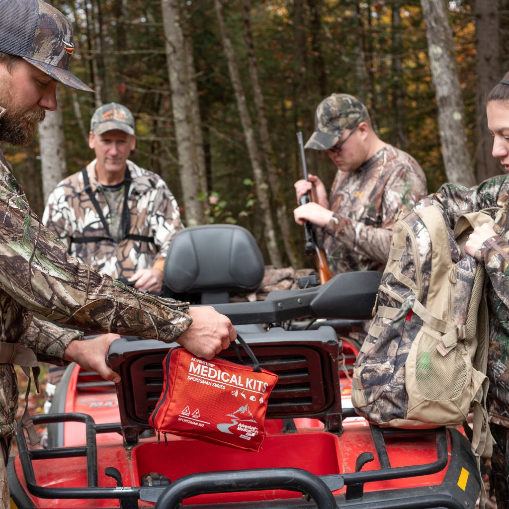 A red medical kit pouch is being placed into a red ATV in a wooded outdoor setting, with several people in camouflage clothing around it.