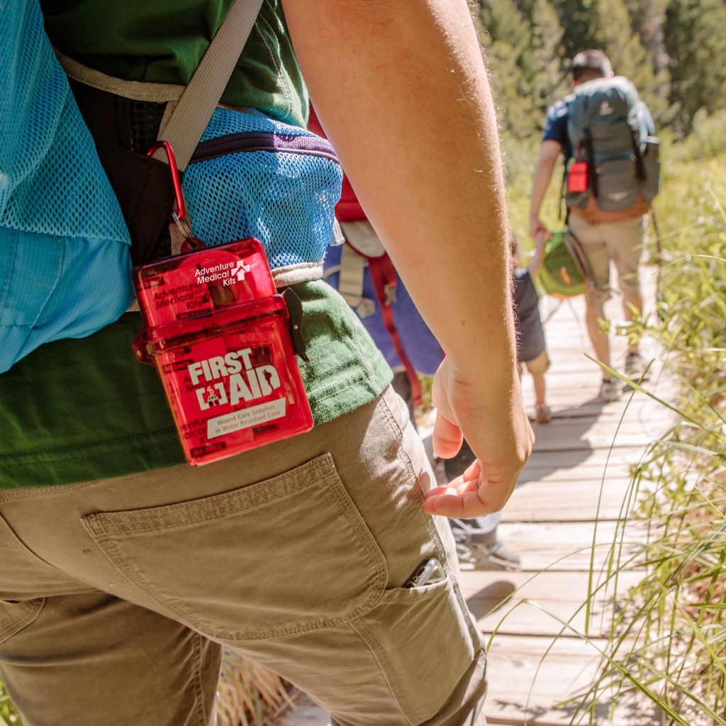 A red first aid kit case clipped to a hiker's waistband outdoors on a wooden trail, with other hikers visible in the background.