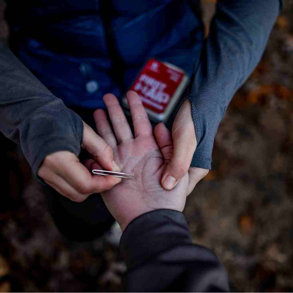 Adventure First Aid, 0.5 Tin person removing splinter from friend's hand with kit in background