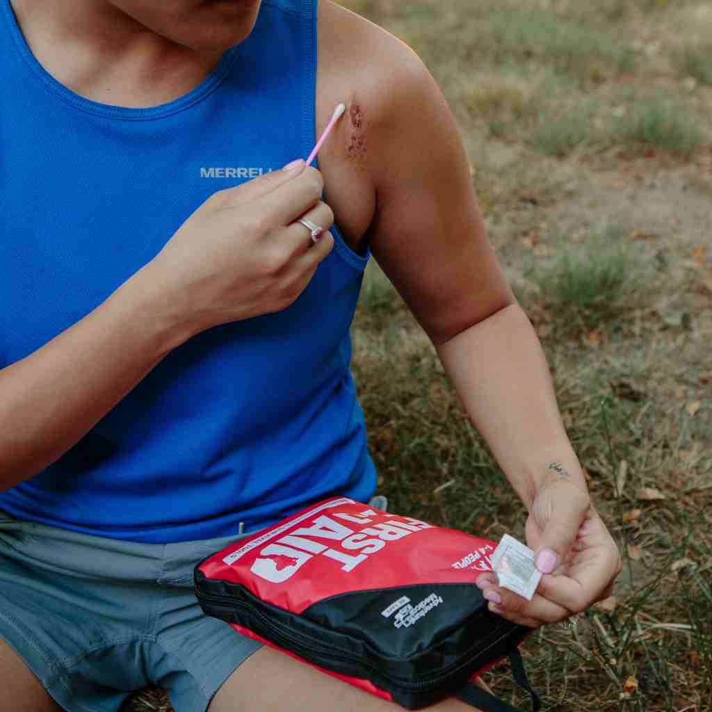 Adventure First Aid, 2.0 person in blue tank top seated using cotton swab to attend to cut on shoulder with kit on lap