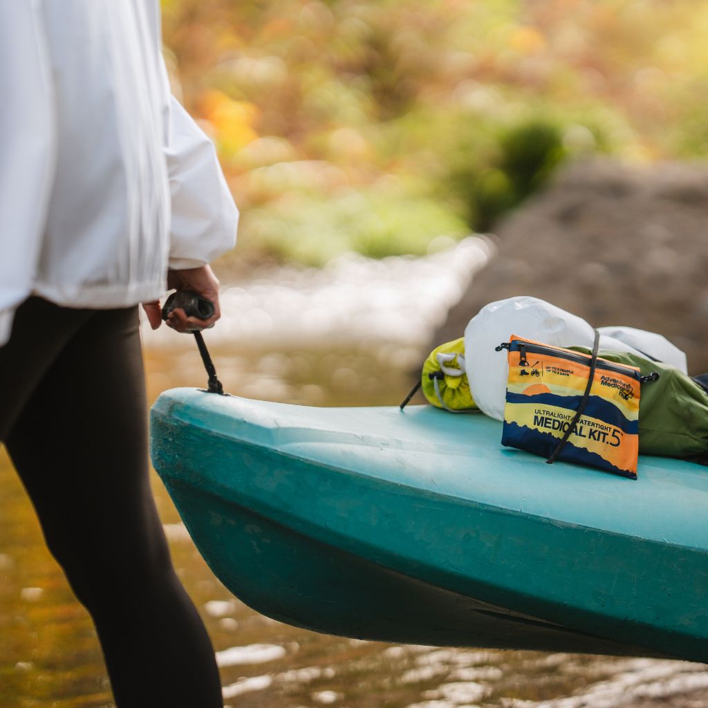 Person holding a paddle with a teal kayak on a riverbank, featuring a medical kit.
