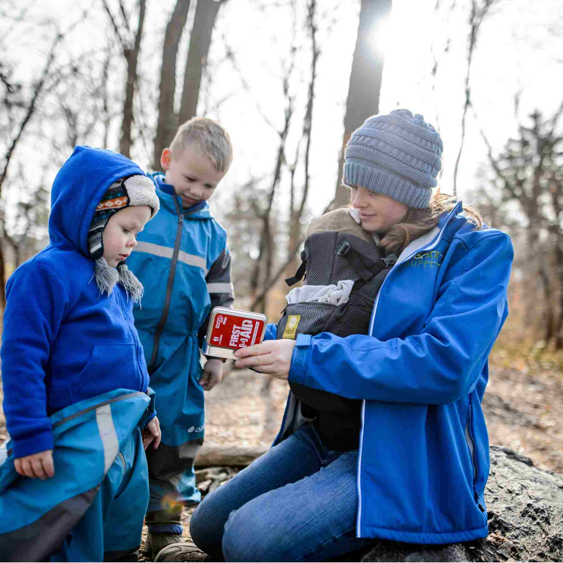 Adventure First Aid, 0.5 Tin woman with baby opening kit in front of two small children all dressed in blue