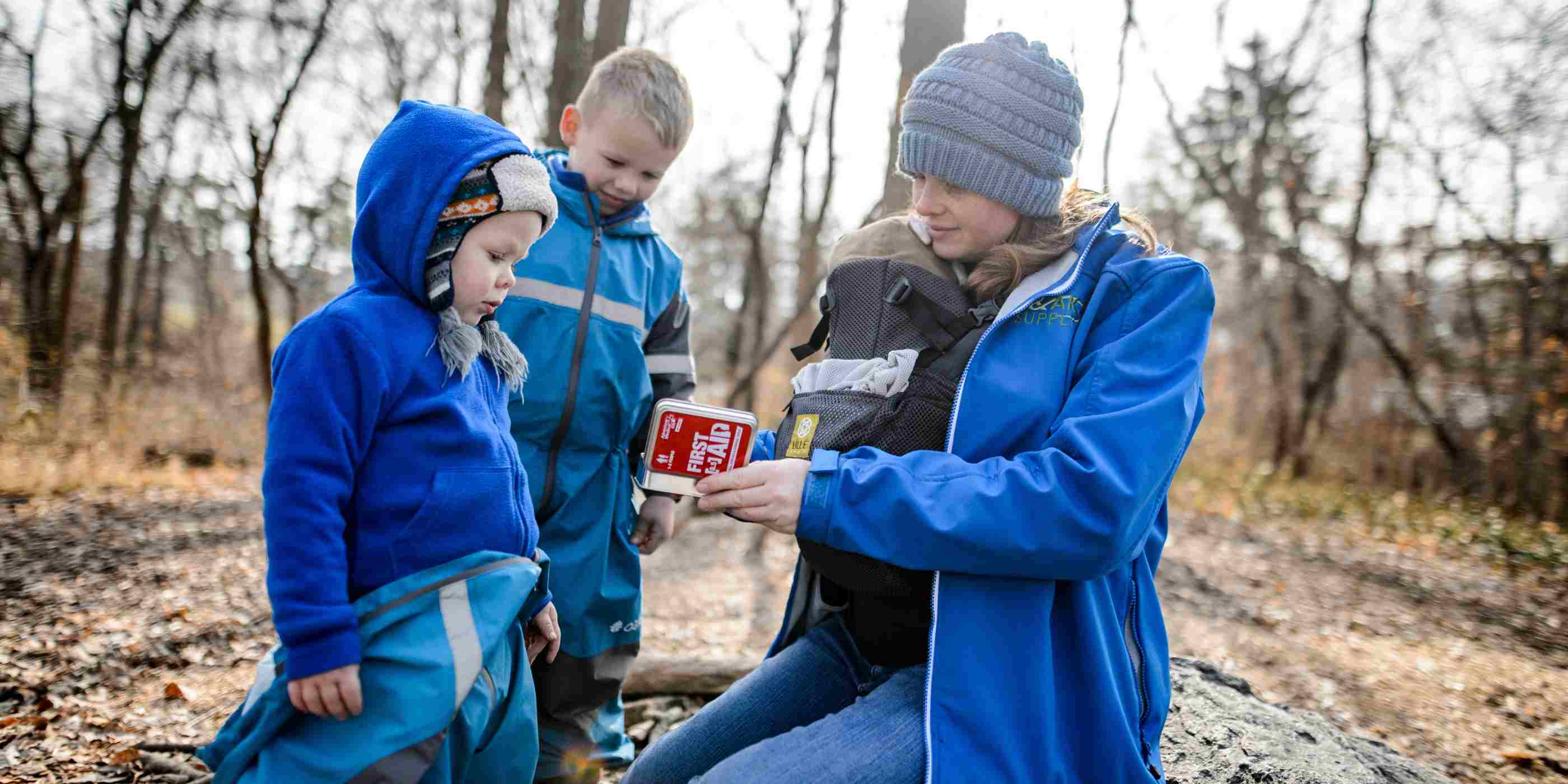 Adventure First Aid, 0.5 Tin woman with baby opening kit in front of two small children all dressed in blue