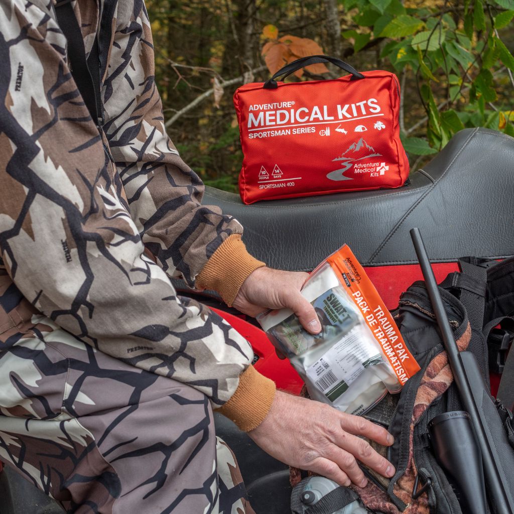 A person in a camouflage jacket packs a medical kit pouch into a backpack on a vehicle outdoors. The red zippered pouch sits on the seat, and the person holds a packaged first aid item.
