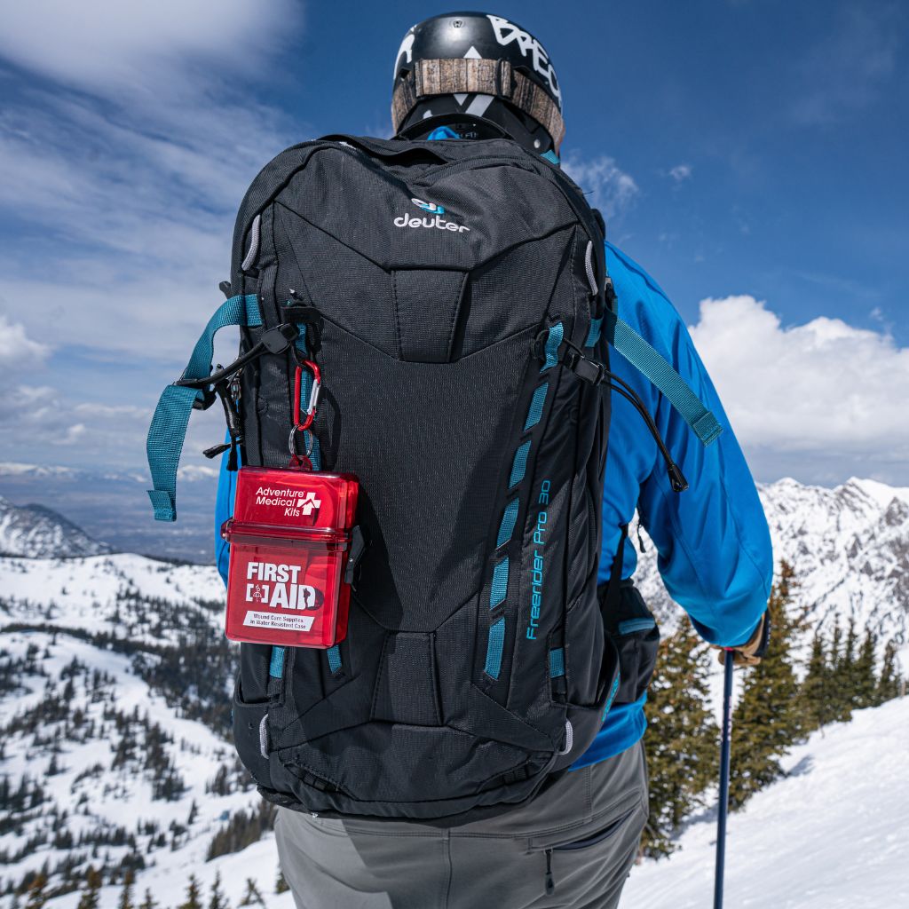 A skier viewed from behind wearing a black Deuter backpack with teal accents, a red first aid pouch, and a ski helmet, set against a snowy mountain landscape under a blue sky.