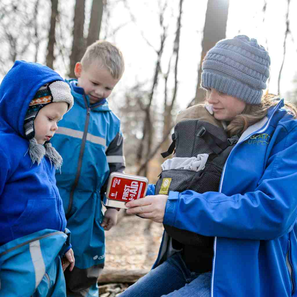 Adventure First Aid, 0.5 Tin woman with baby opening kit in front of two small children all dressed in blue