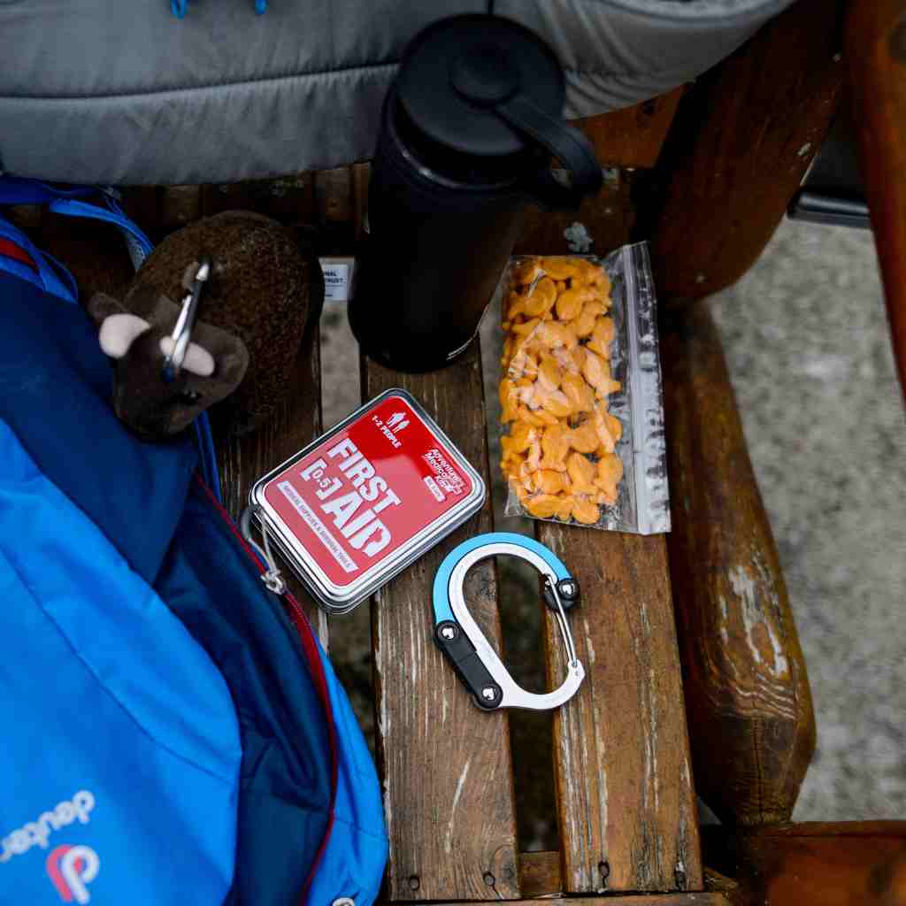 Adventure First Aid, 0.5 Tin kit next to bag of snacks and carabiner on wooden bench