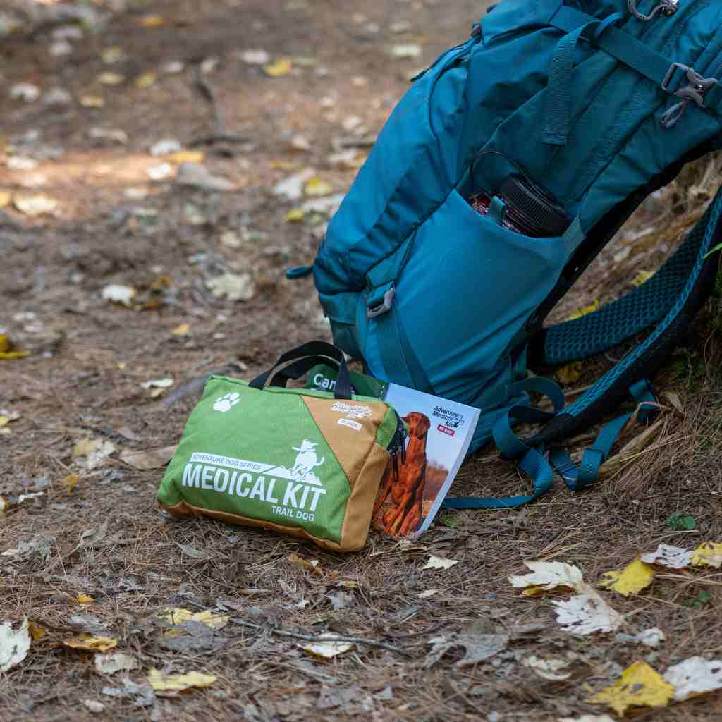 Adventure Dog Medical Kit - Vet in a Box kit and book next to blue backpack on ground