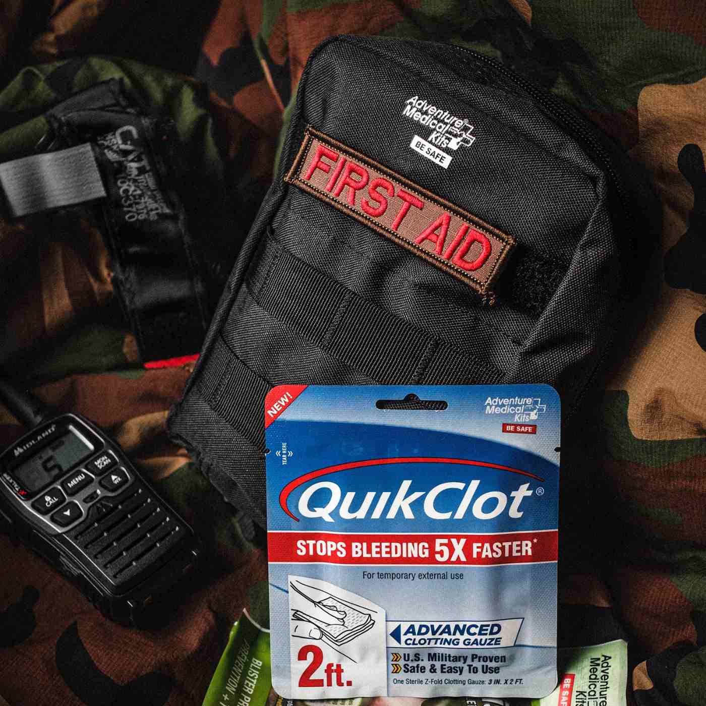 MOLLE Bag Trauma Kit 1.0 - Black with First Aid patch laid out on camo background with radio and QuikClot in the front